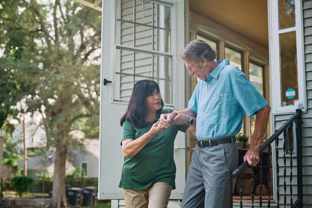 A caregiver in a green shirt helps an elderly man in a blue button-up shirt navigate the front steps of his home, gently holding his hand for support.